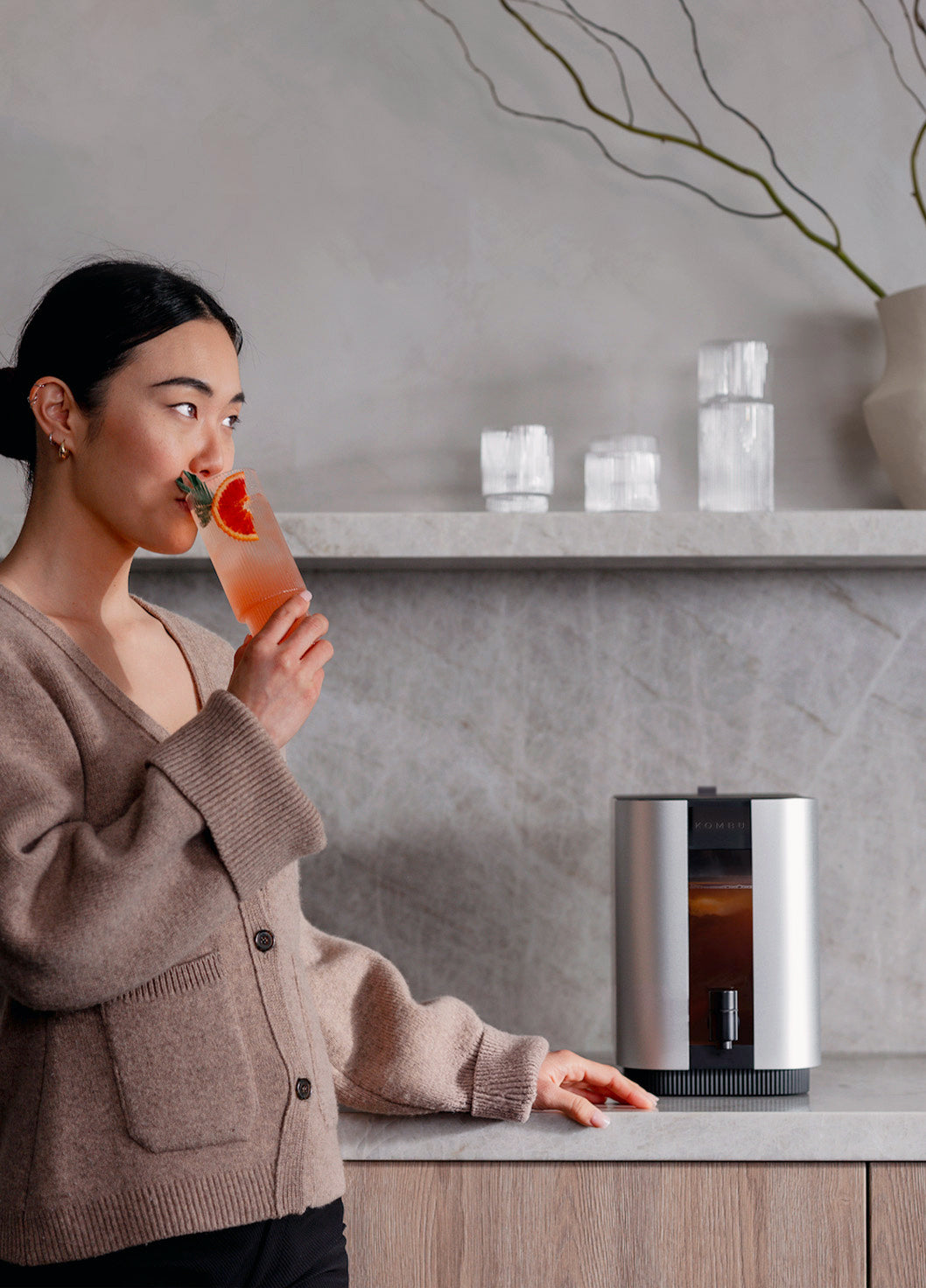 A woman in a beige cardigan stands at a kitchen counter, drinking from a glass with a pink drink and orange slice garnish. A stainless steel coffee machine sits on the counter against a gray stone wall with a shelf in the background.